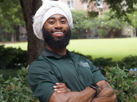 Smiling man with a white headwrap and beard, wearing a Tulane Housing & Residence polo shirt with arms crossed.